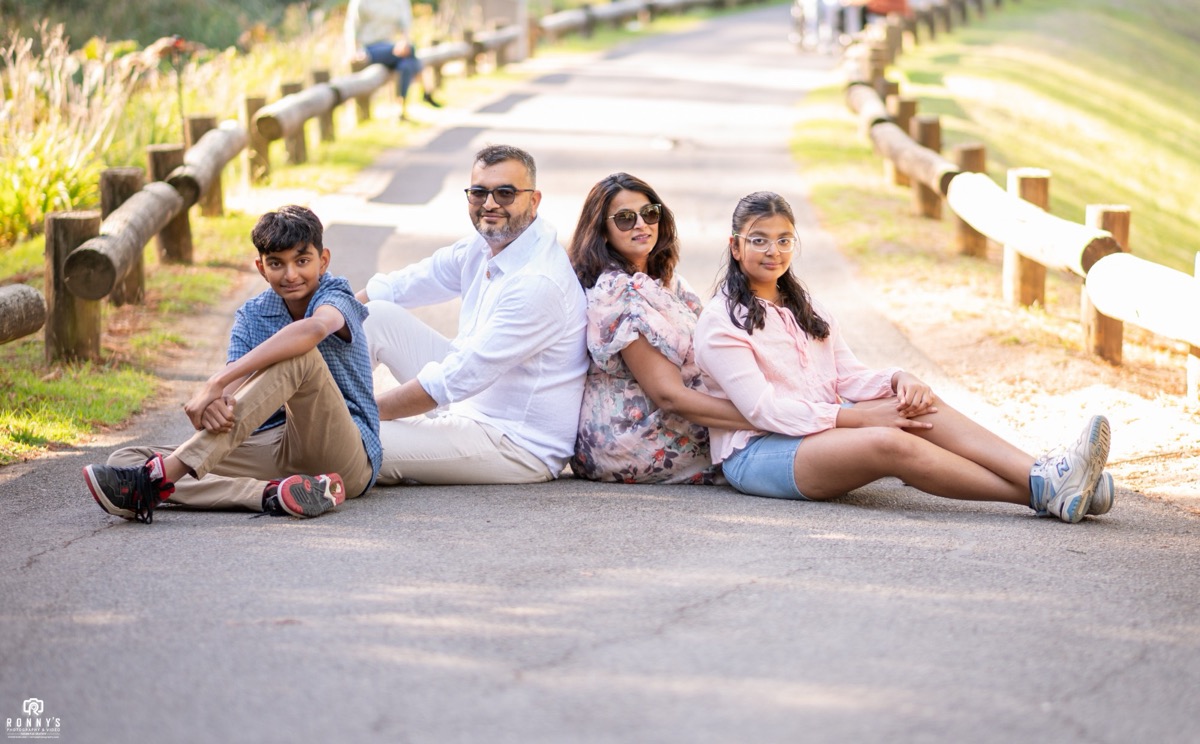 A family of four sitting back to back on a park path in the sunshine